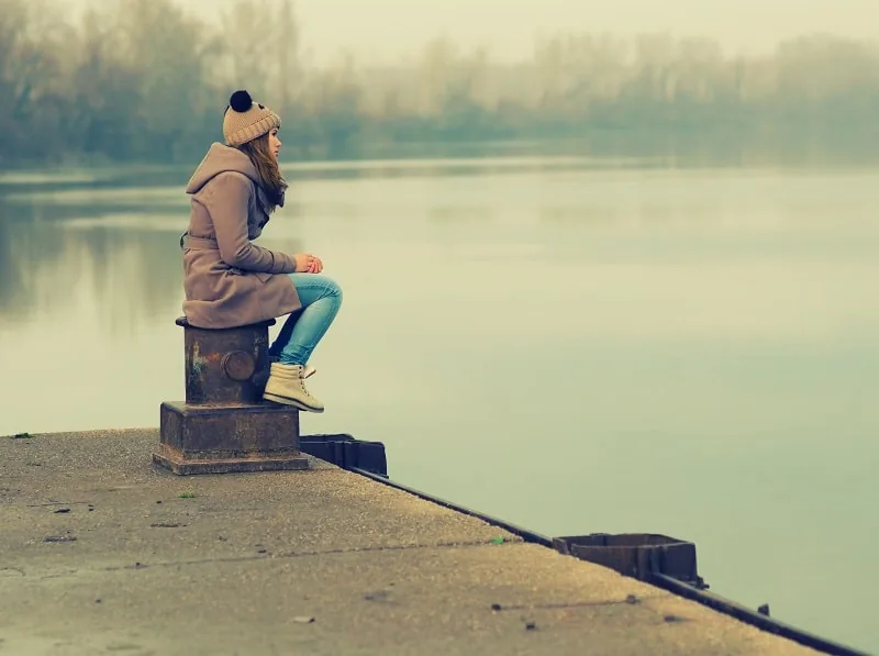 A thoughtful person in a winter coat and knit hat sitting alone by a quiet lake, reflecting during recovery from addiction around the holidays.
