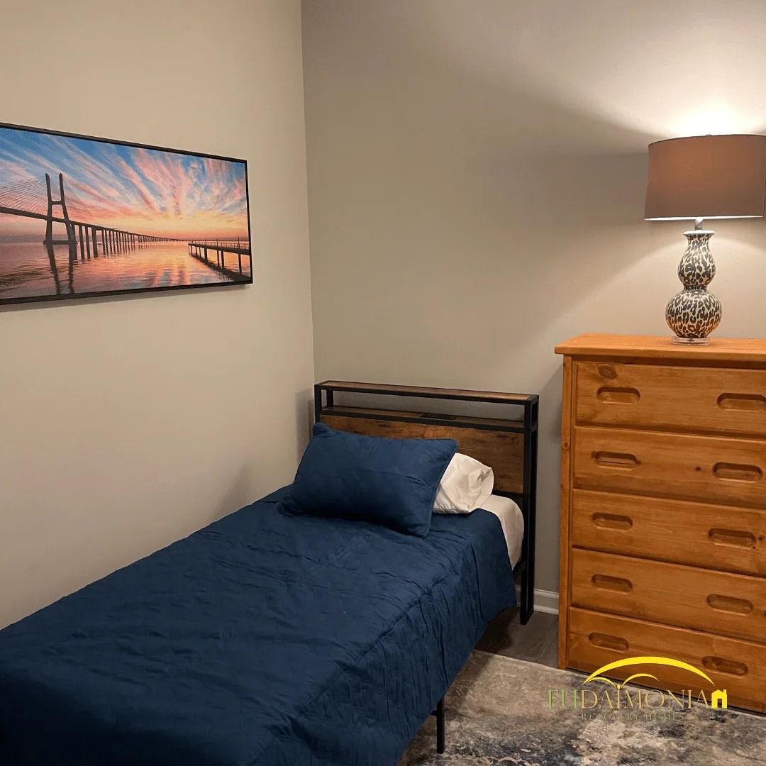 Bedroom with a single bed in blue bedding, a wooden dresser, table lamp and framed bridge photograph.