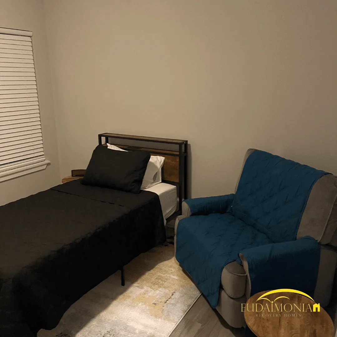 Bedroom with a single bed in dark bedding, a blue upholstered armchair, and a blinded window.