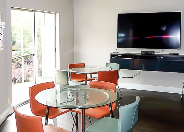 Community room with round glass tables, colorful chairs, a wall-mounted TV, and a sliding glass door.
