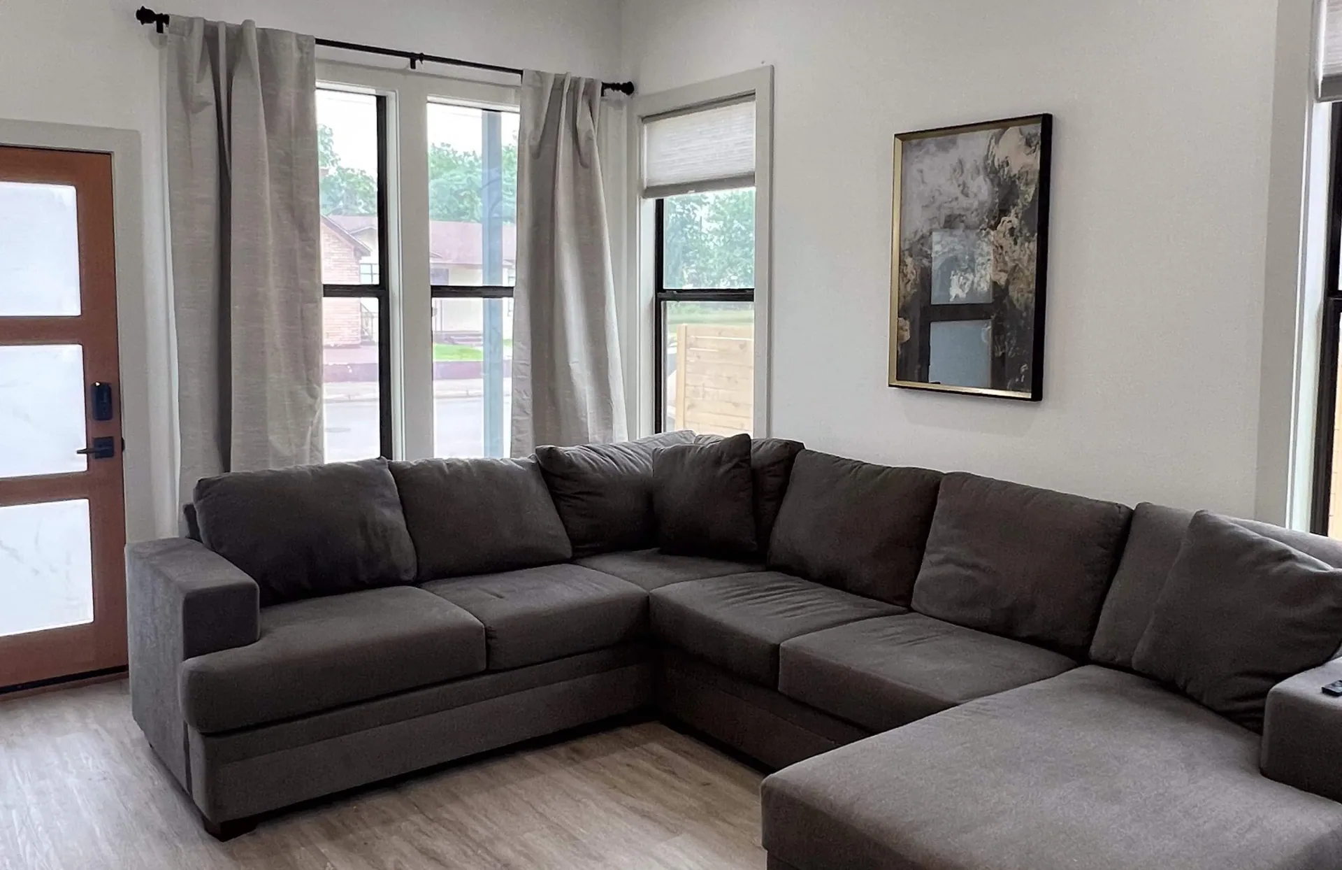 Living room with a large gray corner sofa, tall curtained windows onto the street, and a framed print on the wall.