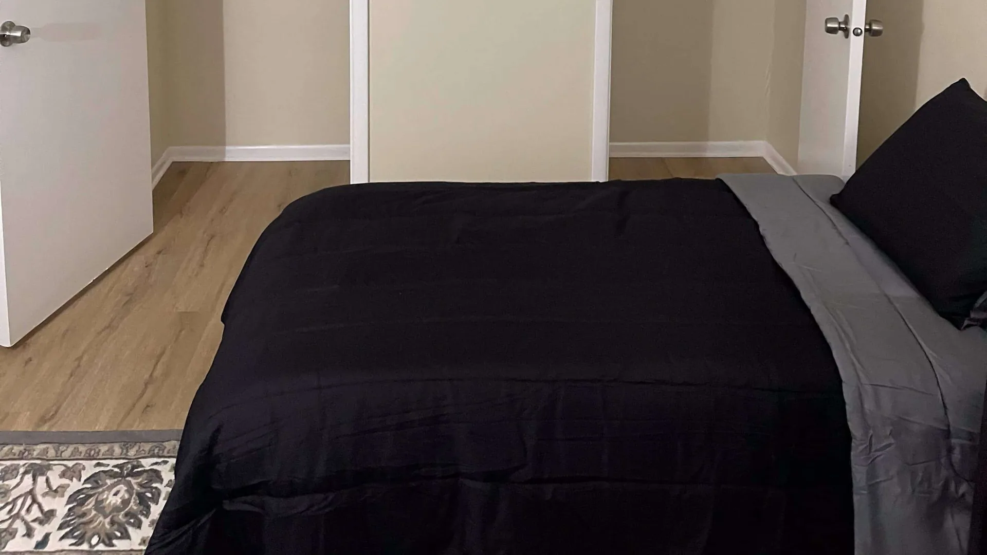 Bedroom with a bed in black bedding against a pale wall, on light wood flooring.