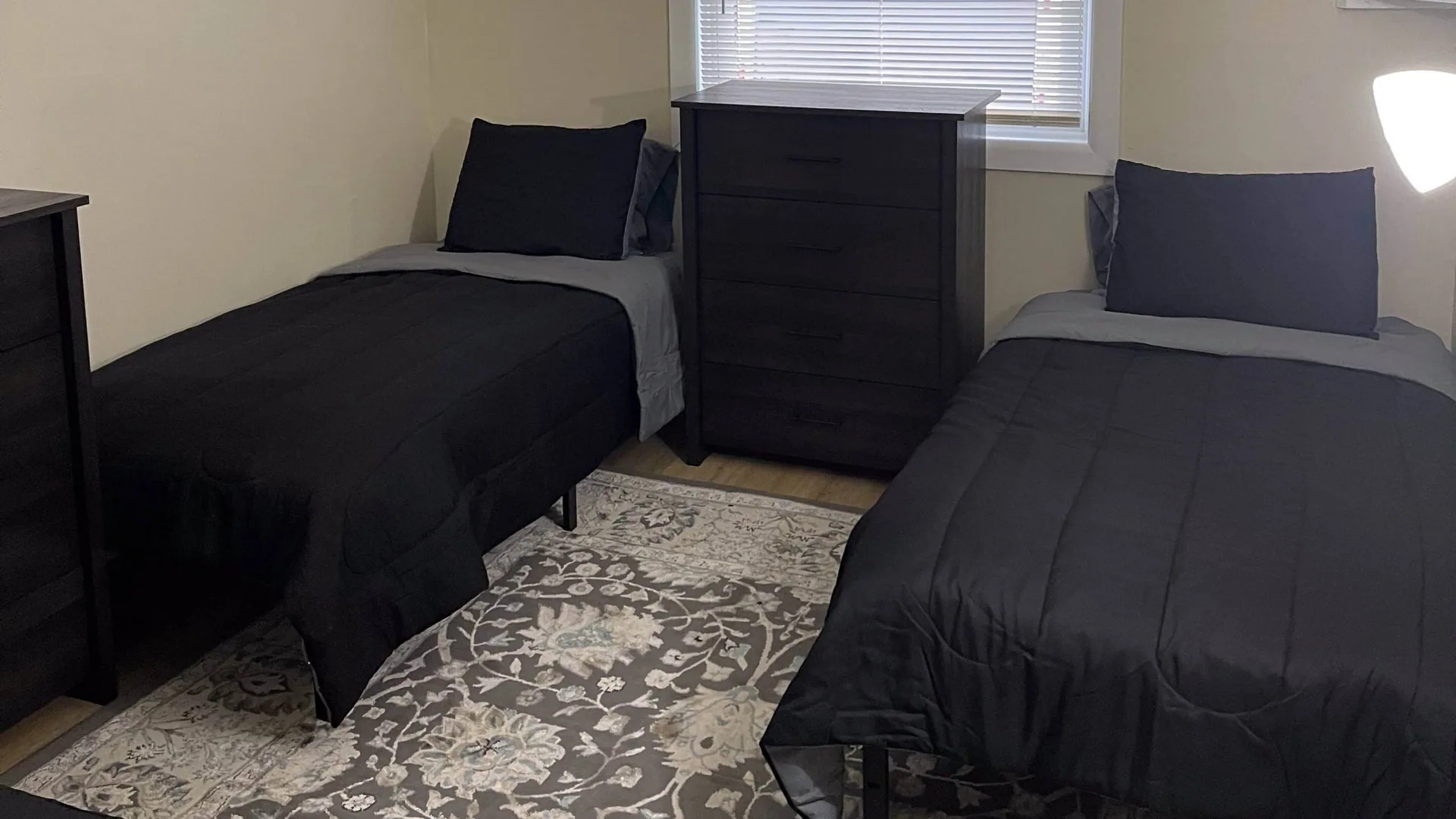 Shared bedroom with two twin beds in black bedding either side of a dark dresser, on a patterned gray rug.