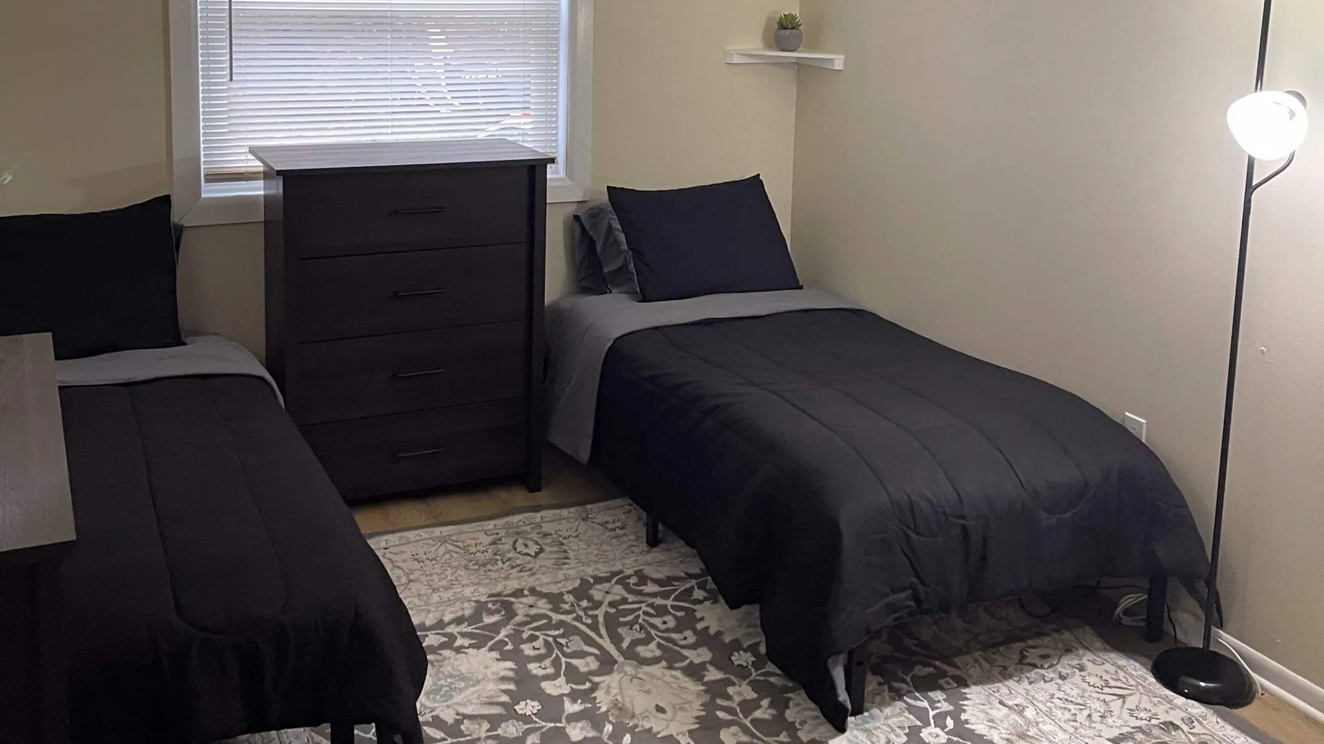 Twin bed in black bedding beside a dark chest of drawers, below a blinded window.