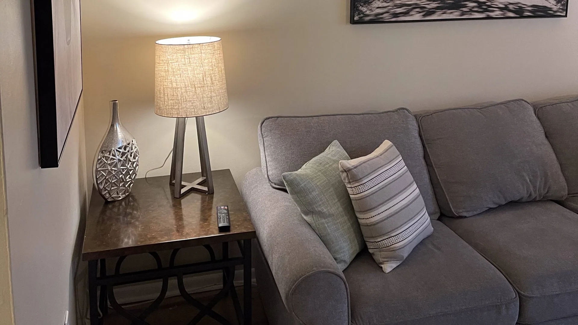 Grey sofa beside a side table with a lamp and vase, under a framed print.