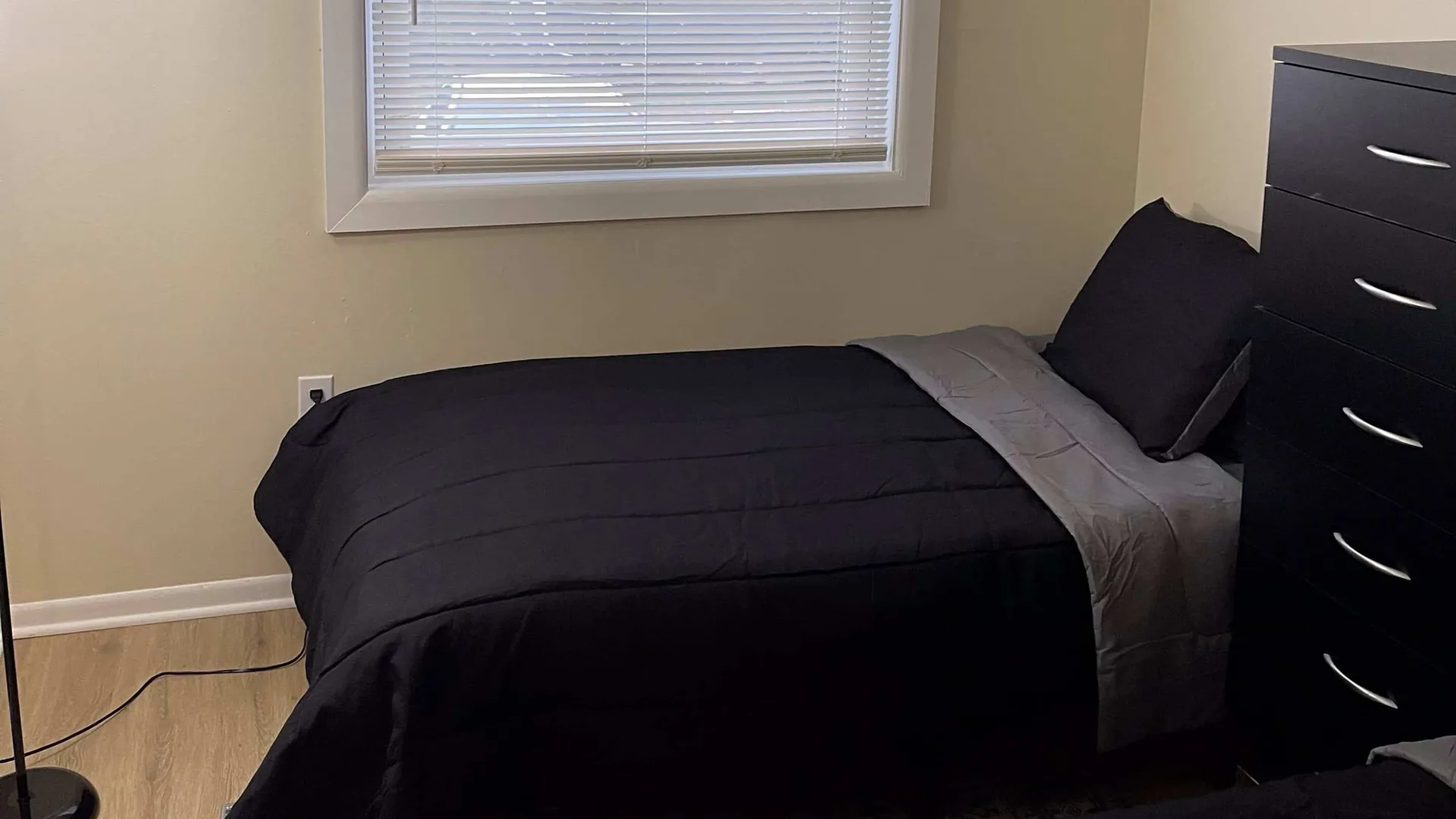 Twin bed in black and gray bedding set against a pale wall by the window.