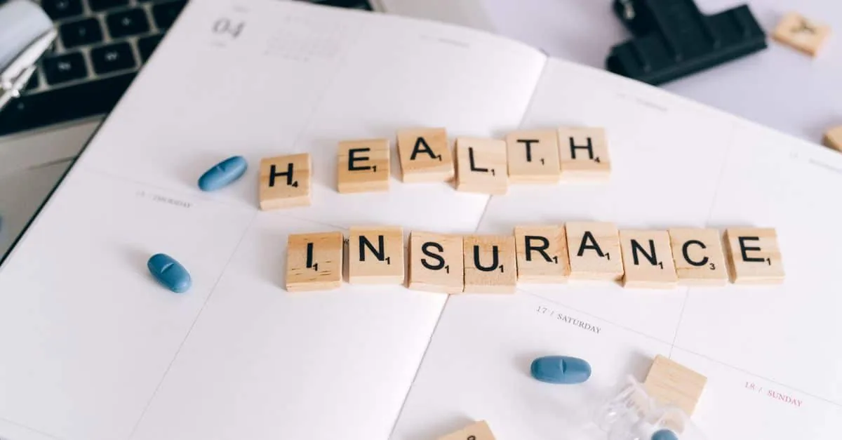 Scrabble tiles spelling 'Health Insurance' on planner with pills and laptop, symbolizing healthcare planning.