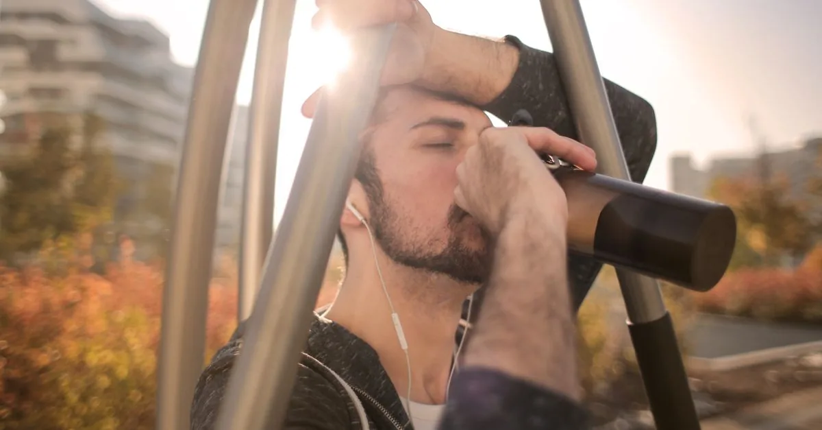 Young thirsty male runner in activewear drinking water from bottle while standing with closed eyes on sports ground and enjoying music in earphones