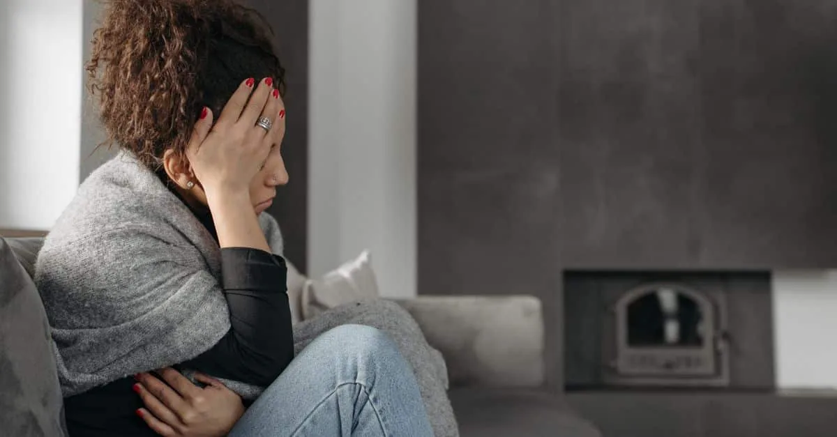 A woman sits on a couch indoors, wrapped in a blanket, holding her head in discomfort.