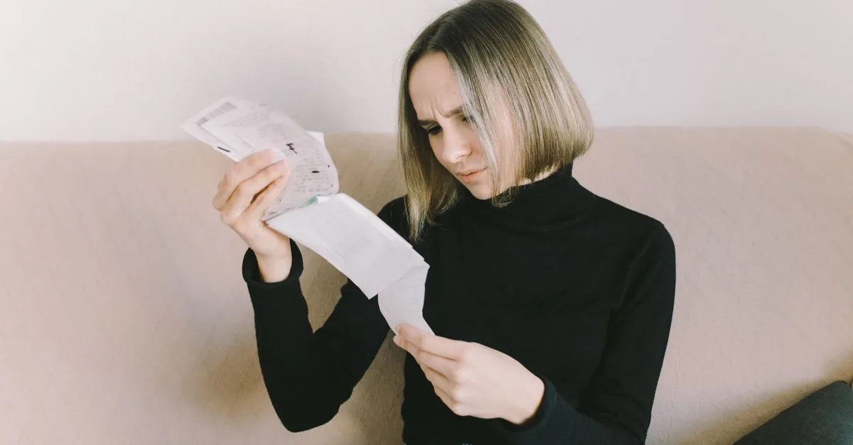 Concerned woman in black sweater examining bills on beige sofa indoors.