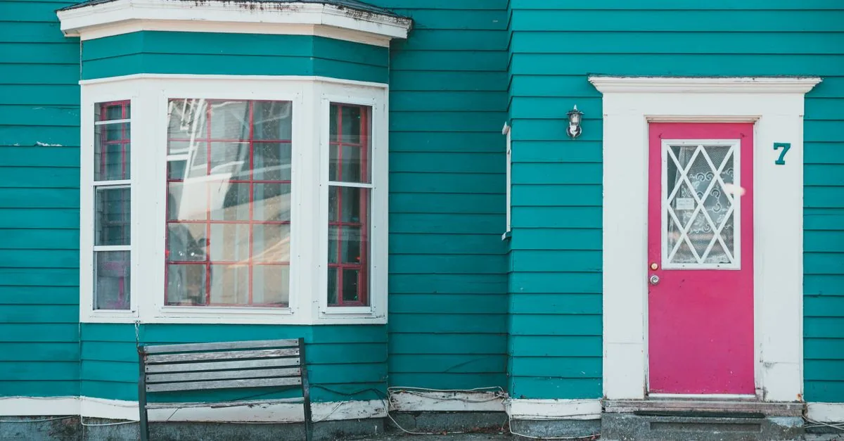 Exterior of a teal house featuring a red door, window, and bench on a sunny day.