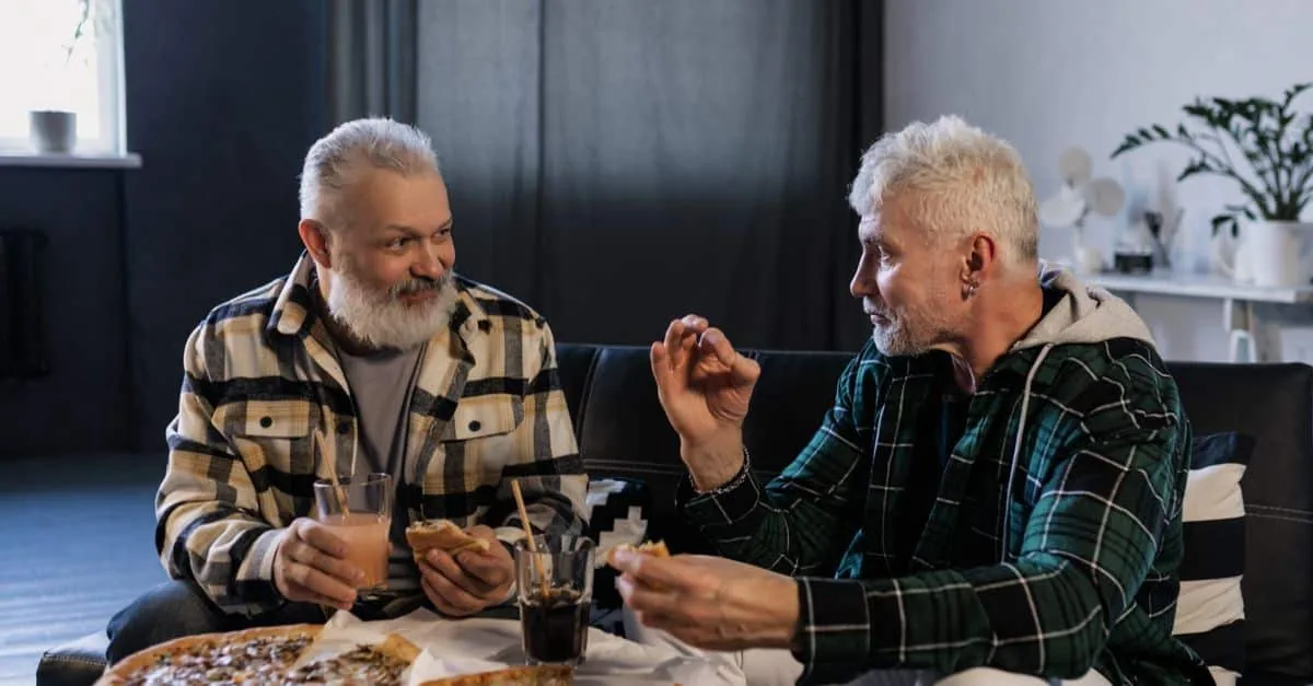 Two senior men enjoying pizza and drinks while chatting in a cozy indoor setting.