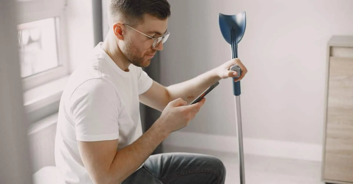 Adult man with eyeglasses using a smartphone indoors, with a crutch beside him. Modern and minimalist setting.