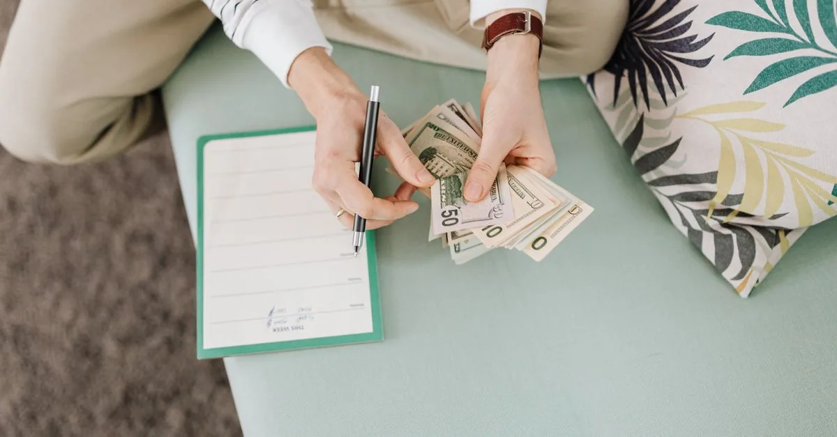 Person holding US dollar bills with a notepad and pen on a soft surface indoors.