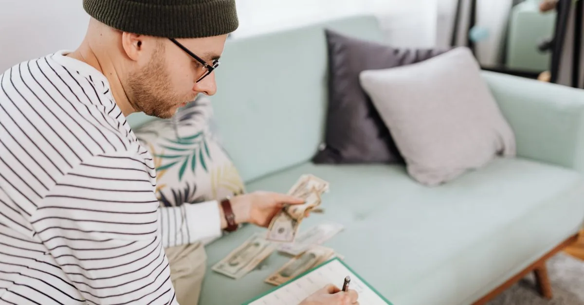 Adult man with beanie counting cash and making notes on a couch.