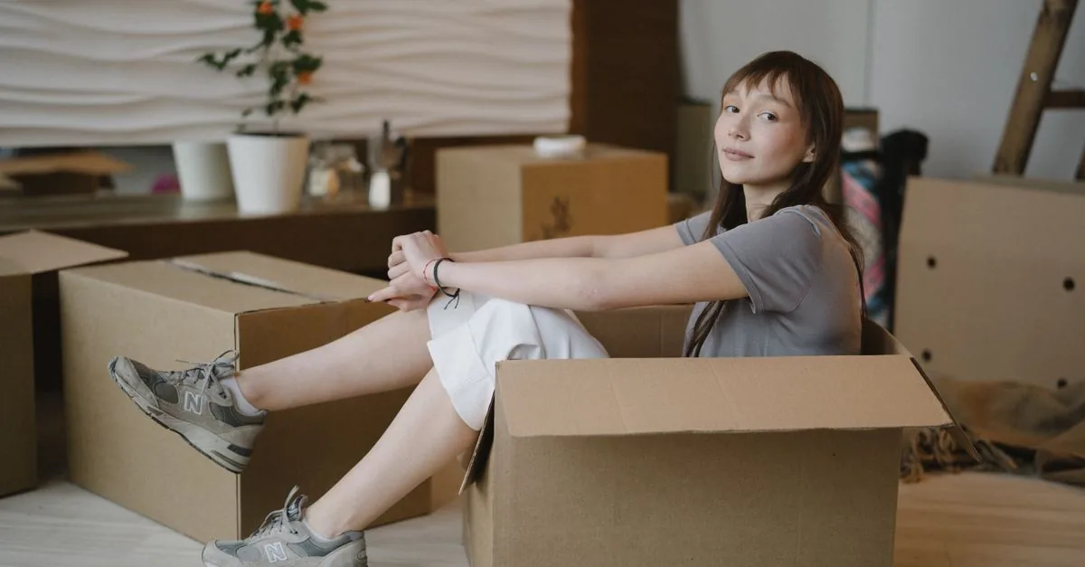 Woman sits in a cardboard box surrounded by moving boxes, symbolizing a new home journey.