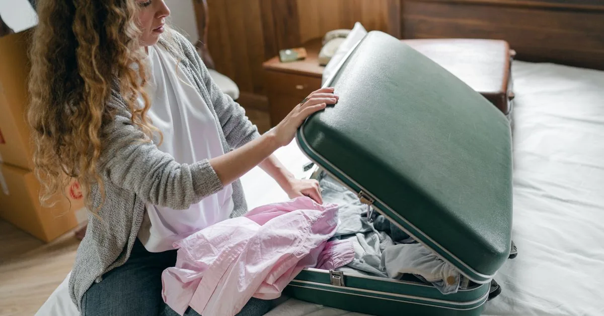 From above concentrated young female in casual wear with long curly hair sitting on bed and packing suitcase before trip