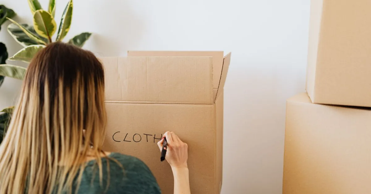 Back view anonymous female in casual outfit writing on cardboard container word cloth while packing belongings and preparing to move