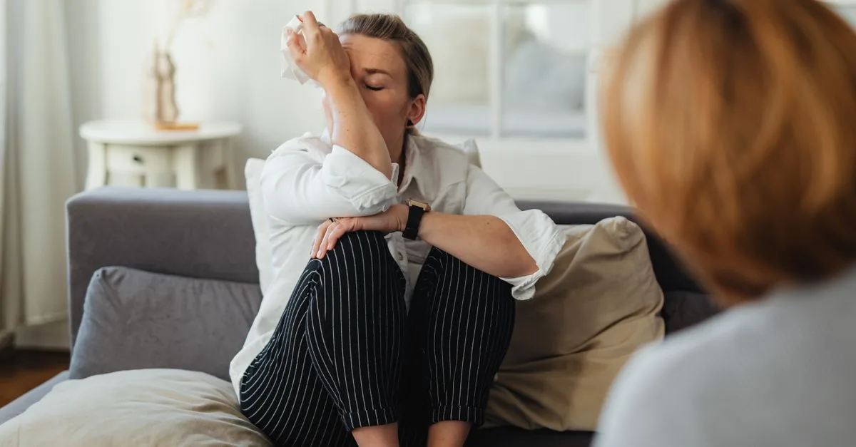 A woman wipes tears during a therapy session on a couch indoors.