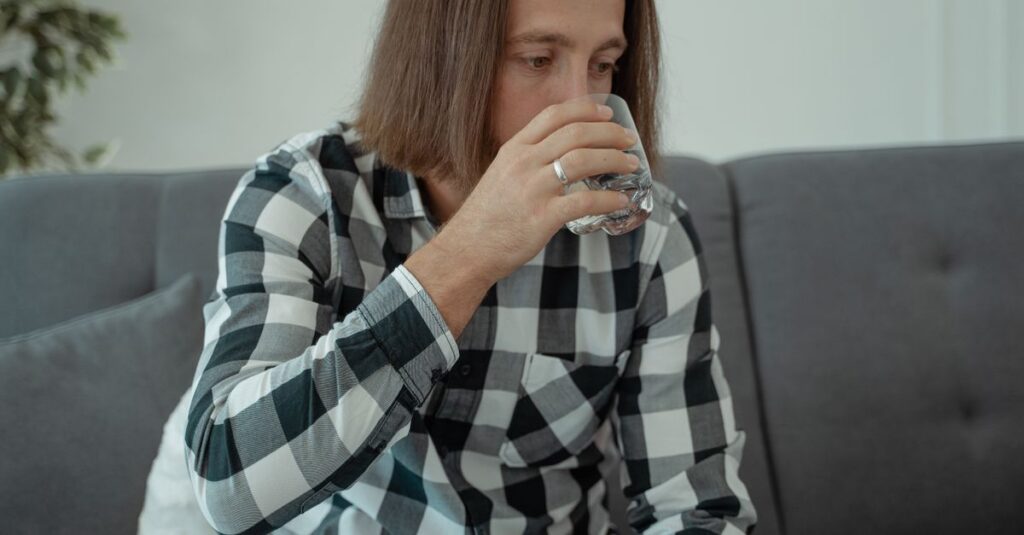 A man with long hair wears a checkered shirt, sitting on a couch, drinking water.