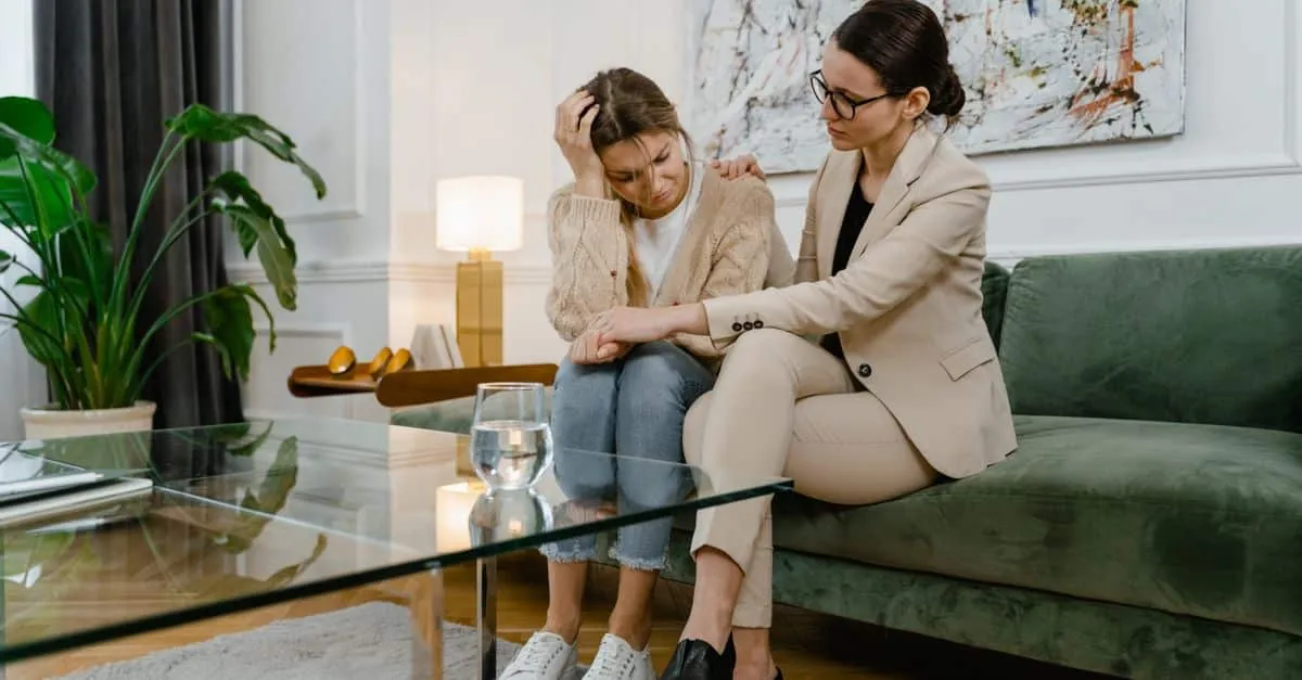 Two women in a living room, one comforting the other on a green sofa.