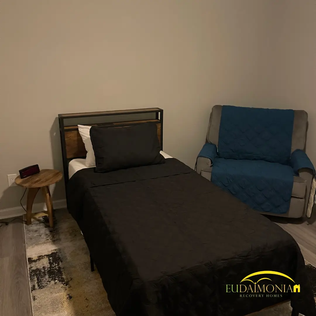 Single bed with dark bedding beside a blue upholstered armchair.