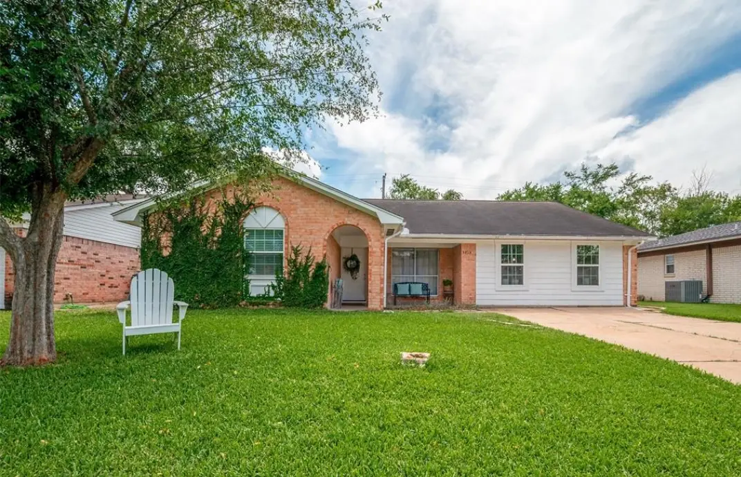 Front of the single-story brick house with a lawn and patio chairs.
