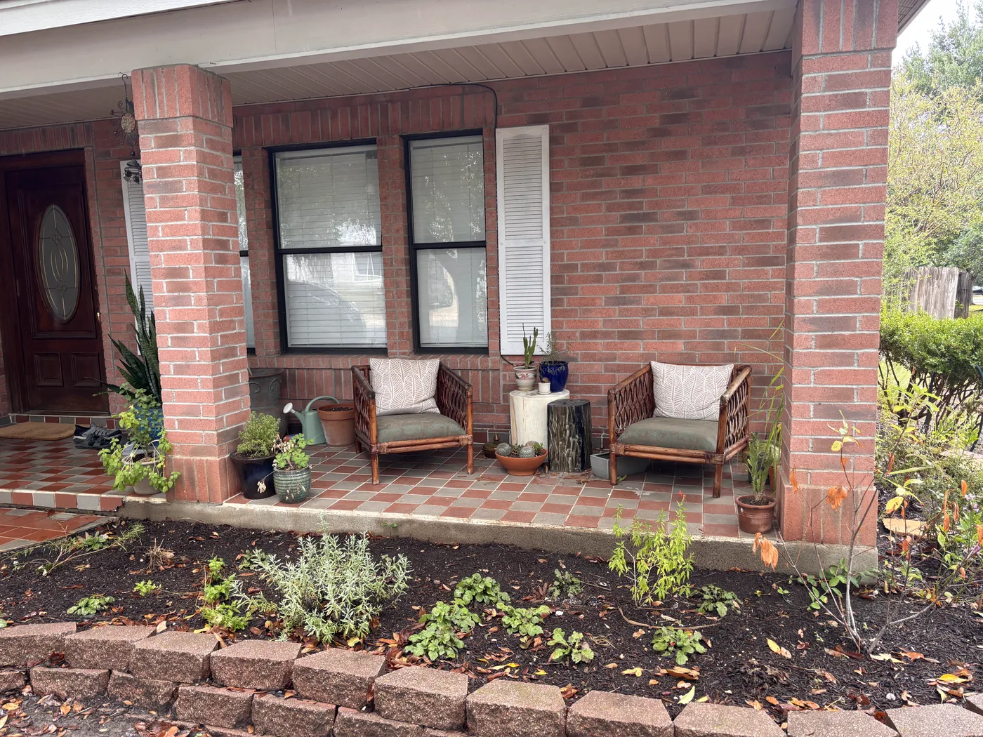Covered brick front porch with two rattan chairs, potted plants and a dark wood front door.