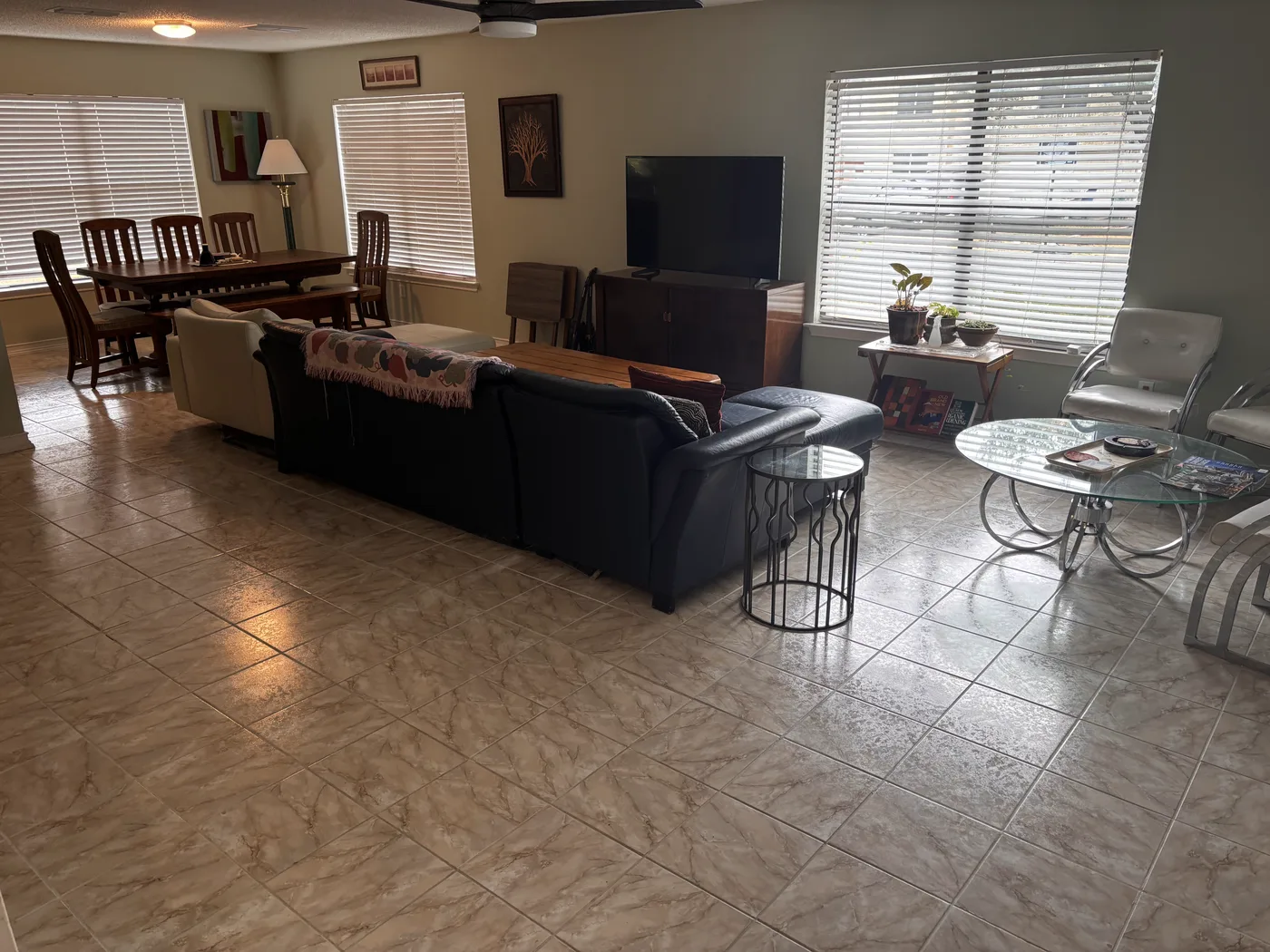 Open living and dining area with a leather sectional, glass tables and a tiled floor.