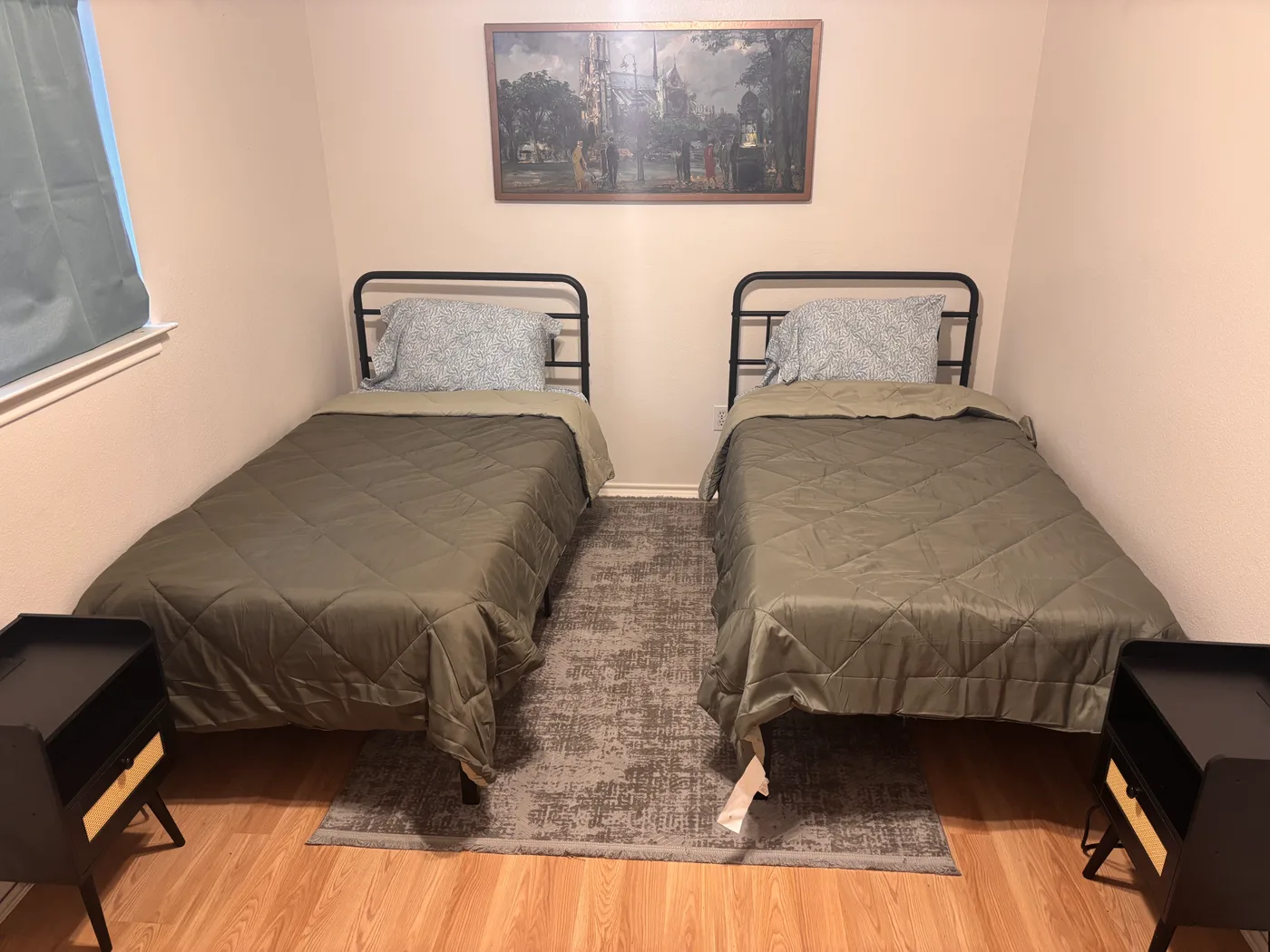 Two beds with black cane nightstands either side, under a framed street painting.