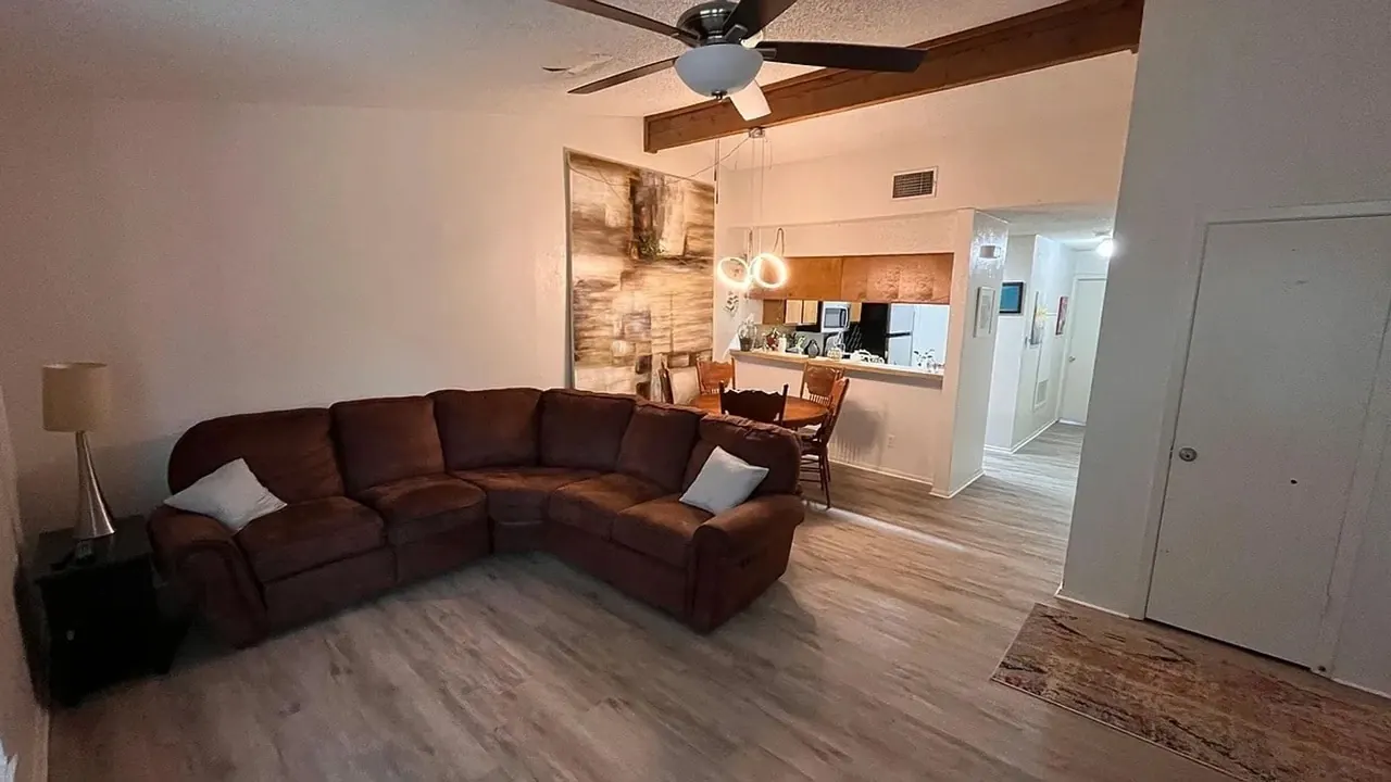 Living area in the South Austin apartment: a brown sectional sofa on wood-look flooring, with a ceiling fan, exposed beam and the kitchen beyond.